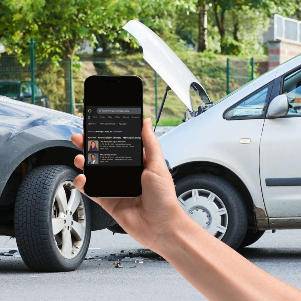 decorate image of a person's hand holding a phone in front of a car crash, with the screen displaying a locally specific attorney google search.